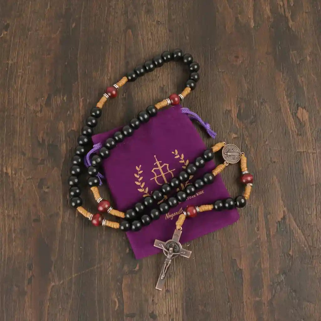 Black rosary beads with wooden and red accents on a purple pouch with a cross symbol, all on a wooden surface.