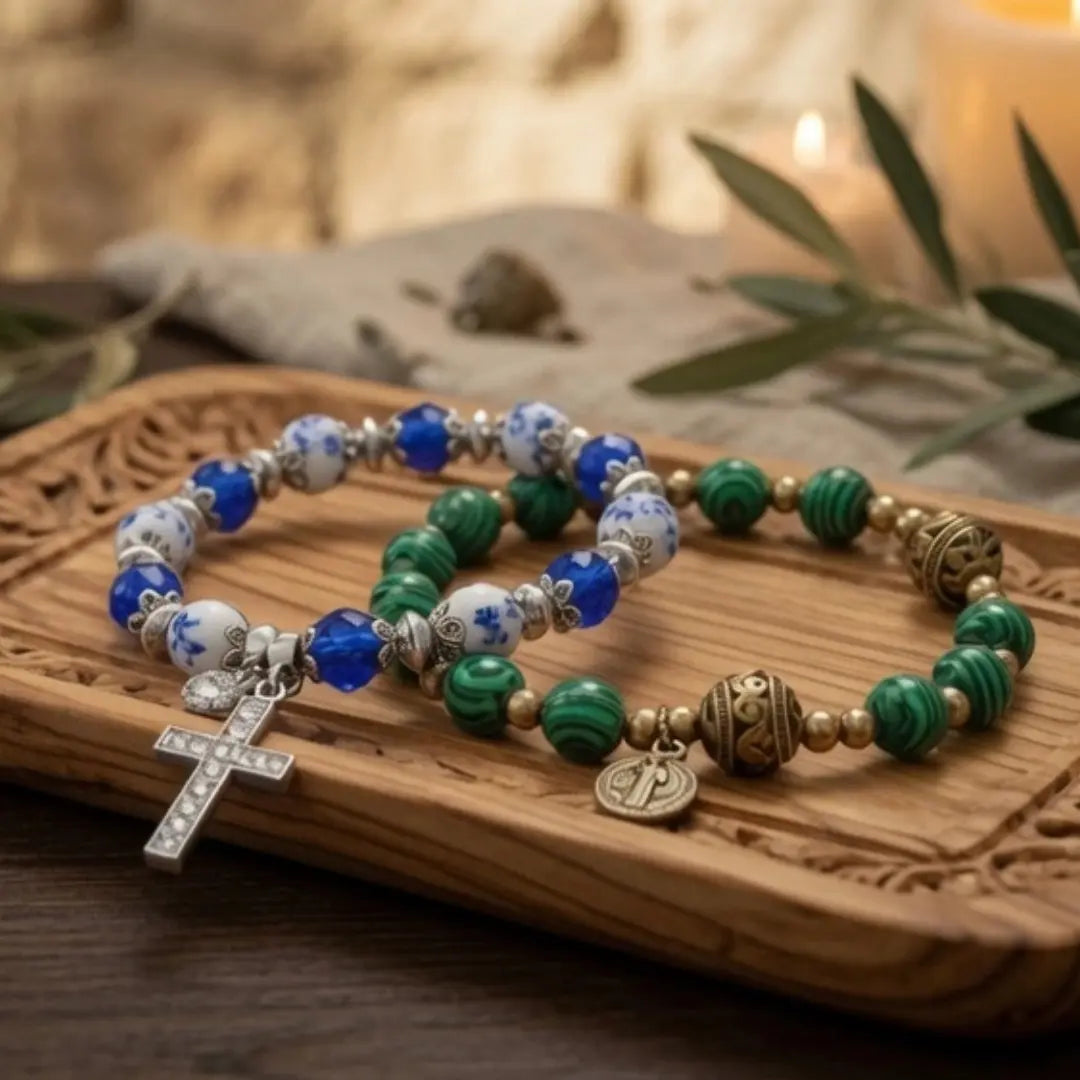 Two beaded catholic bracelets with blue and white floral beads, green malachite beads, gold-toned spacer beads, and a silver cross charm, displayed on a carved wooden tray with candles in the background.