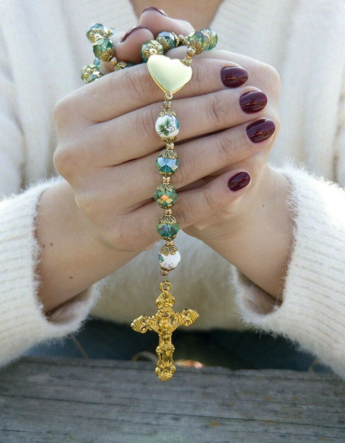 Hands holding a rosary with a gold cross, wearing a white sweater.